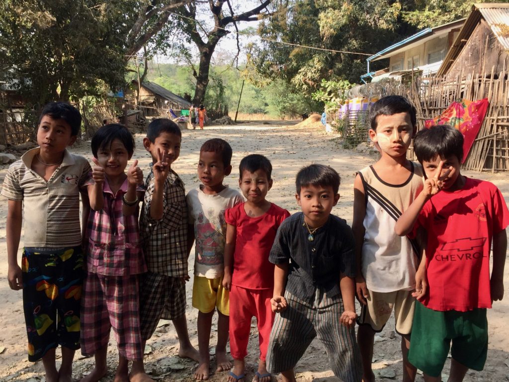 Eight kids in line posing for a group photo. Dirt road and bamboo homes in the background.