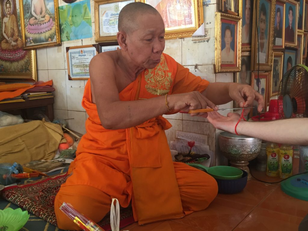 Orange robed Buddhist monk with scissors trimming the excess from an orange and red bracelet he has just woven onto my wrist
