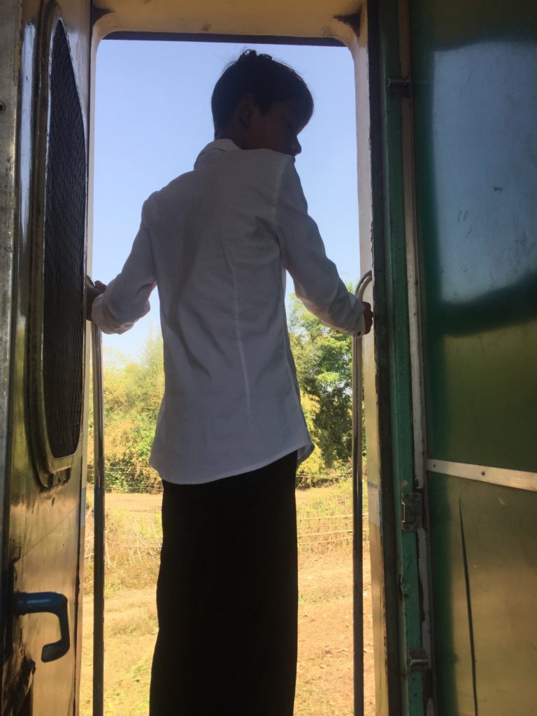 Young man stands in the doorway and looks out at the countryside on the train to Mawlamyine